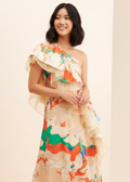 A woman modeling a one-shoulder ruffled maxi dress with a colorful abstract floral print on a cream base, set against a plain beige background.