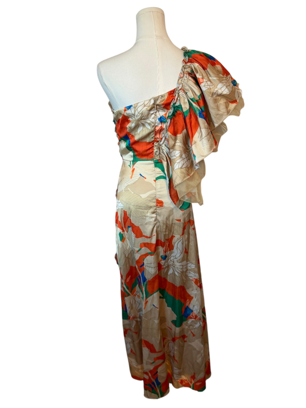 A one-shoulder maxi dress with a draped, ruffled neckline, featuring an abstract floral pattern in orange, green, blue, and white on a beige background, displayed on a mannequin.
