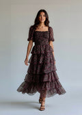 A woman modeling a dark floral tiered midi dress with puff sleeves and a square neckline, standing against a plain light gray studio background.