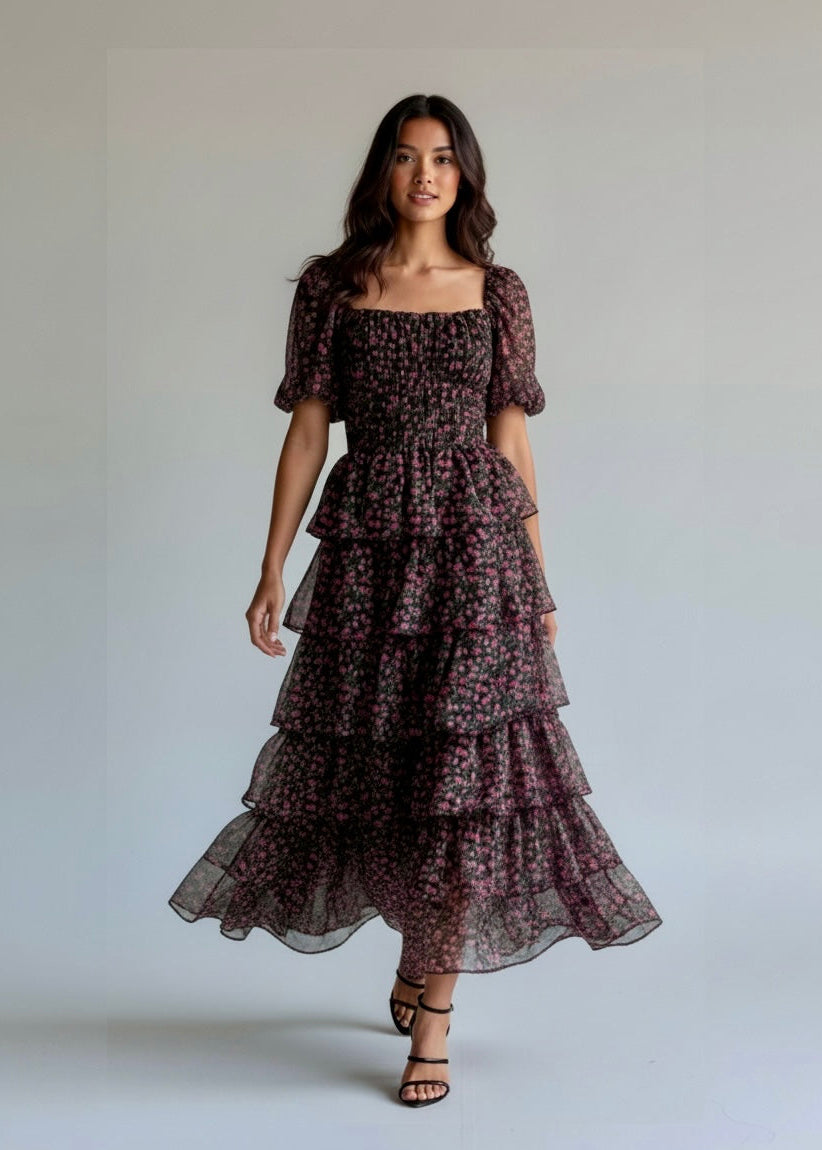 A woman modeling a dark floral tiered midi dress with puff sleeves and a square neckline, standing against a plain light gray studio background.