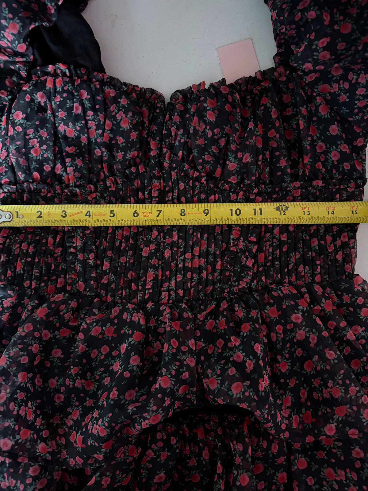 A close-up of a black floral dress with red and pink flowers, featuring a smocked bodice and ruffled skirt, with a yellow tape measure showing the waist measurement.